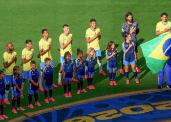 Brazil national team players and children stand in line with hands over their hearts on a soccer pitch, a flag bearer holding the Brazilian flag nearby