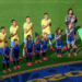 Brazil national team players and children stand in line with hands over their hearts on a soccer pitch, a flag bearer holding the Brazilian flag nearby