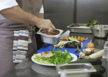 Chef in apron sauces a plate of greens and salmon, with a metal tray in hand and vegetables on a blue cutting board nearby
