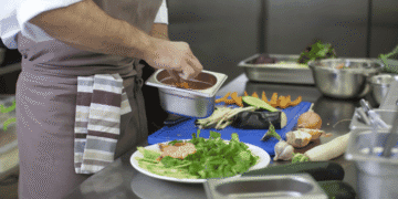 Chef in apron sauces a plate of greens and salmon, with a metal tray in hand and vegetables on a blue cutting board nearby