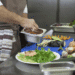 Japão congela novas autorizações para estrangeiros em restaurantes e expõe pressão sobre a mão de obra - ThingsOut Chef in apron sauces a plate of greens and salmon, with a metal tray in hand and vegetables on a blue cutting board nearby