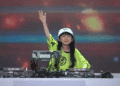 Young female DJ in a lime-green shirt and white cap raises her hand while performing behind a mixer labeled '2S MUSIC' on a stage with colorful lights.