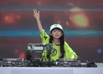 Young female DJ in a lime-green shirt and white cap raises her hand while performing behind a mixer labeled '2S MUSIC' on a stage with colorful lights.