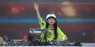 Japonesa de 9 anos se destaca entre os DJs mais jovens do mundo - ThingsOut Young female DJ in a lime-green shirt and white cap raises her hand while performing behind a mixer labeled '2S MUSIC' on a stage with colorful lights.