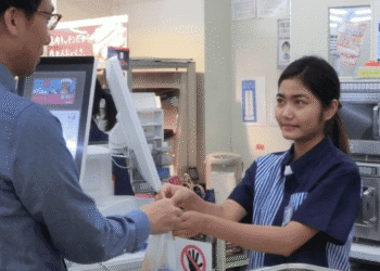 Cashier in a blue uniform hands a plastic bag to a customer at the checkout counter.