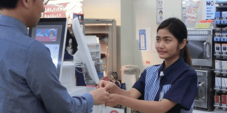 Cashier in a blue uniform hands a plastic bag to a customer at the checkout counter.