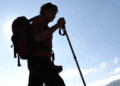 Silhouette of a hiker with a red backpack and trekking pole against a bright blue sky at daytime.