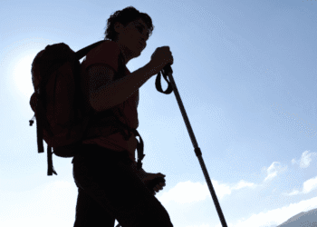 Silhouette of a hiker with a red backpack and trekking pole against a bright blue sky at daytime.