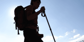 Silhouette of a hiker with a red backpack and trekking pole against a bright blue sky at daytime.