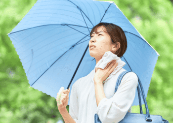 Woman holding a large blue umbrella in a park, wiping sweat from her neck with a towel under bright greenery.