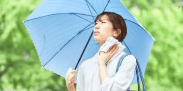 Woman holding a large blue umbrella in a park, wiping sweat from her neck with a towel under bright greenery.