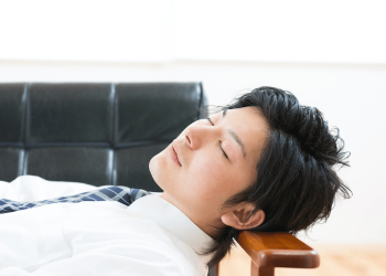 Man in a white shirt and tie resting with his head on an armchair, eyes closed.