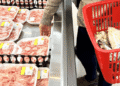 Iene fraco pressiona preços e leva carne importada a recorde no Japão - ThingsOut Person in a grocery store picking up trays of pink meat from a cold case while a red shopping basket sits nearby filled with packaged goods, shopper wearing a sweater and jeans.