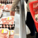 Person in a grocery store picking up trays of pink meat from a cold case while a red shopping basket sits nearby filled with packaged goods, shopper wearing a sweater and jeans.