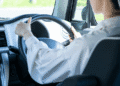 Person driving a car, hands on the steering wheel, dashboard and interior visible from the passenger side window behind the driver