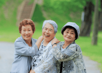 Three elderly women smiling and embracing each other in a green park.