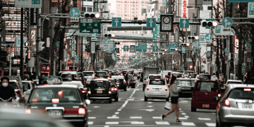 Motoristas no Japão terão novas regras de velocidade a partir de setembro - ThingsOut Busy Japanese city street with cars, pedestrians, and numerous green overhead traffic signs against tall buildings.