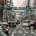 Busy Japanese city street with cars, pedestrians, and numerous green overhead traffic signs against tall buildings.