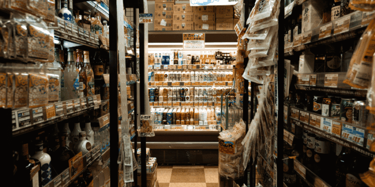 Petróleo caro pressiona custos e pode acelerar reajustes de preços no Japão - ThingsOut A narrow supermarket aisle with shelves stocked full of bottled drinks and a refrigerated section at the end.