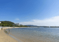 A calm riverside beach with people strolling along the shore and calm water, hills and buildings in the background under a clear blue sky.