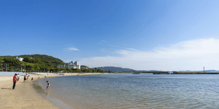 A calm riverside beach with people strolling along the shore and calm water, hills and buildings in the background under a clear blue sky.