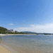 A calm riverside beach with people strolling along the shore and calm water, hills and buildings in the background under a clear blue sky.