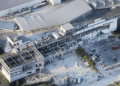 Aerial view of a damaged mall with collapsed roof and debris scattered in the parking lot, AEON Mall signage visible on the building path nearby.