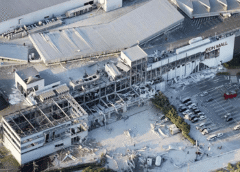 Aerial view of a damaged mall with collapsed roof and debris scattered in the parking lot, AEON Mall signage visible on the building path nearby.