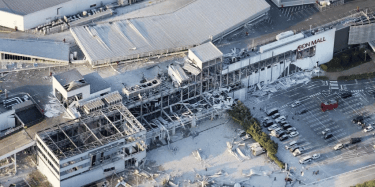 Aerial view of a damaged mall with collapsed roof and debris scattered in the parking lot, AEON Mall signage visible on the building path nearby.